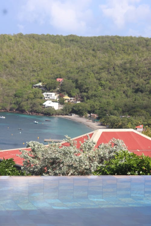 Vue panoramique de la baie tropicale depuis la piscine de l'hôtel INHÉRENCE avec bâtiments aux toits rouges, eau turquoise et forêt luxuriante à la Martinique