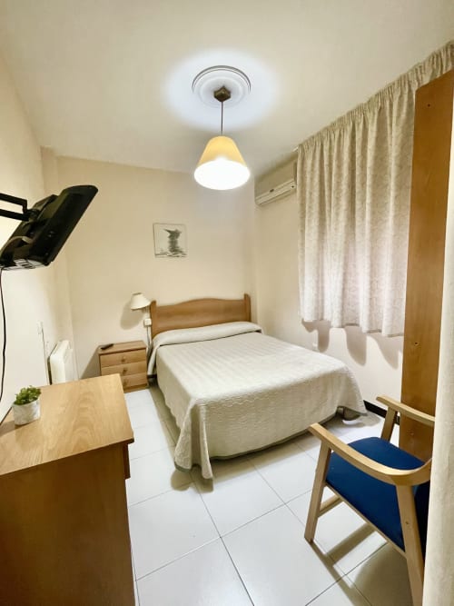 Habitación individual en Hotel JM El Emigrante con cama de madera, escritorio, silla, lámpara colgante y vista al patio interior desde la ventana