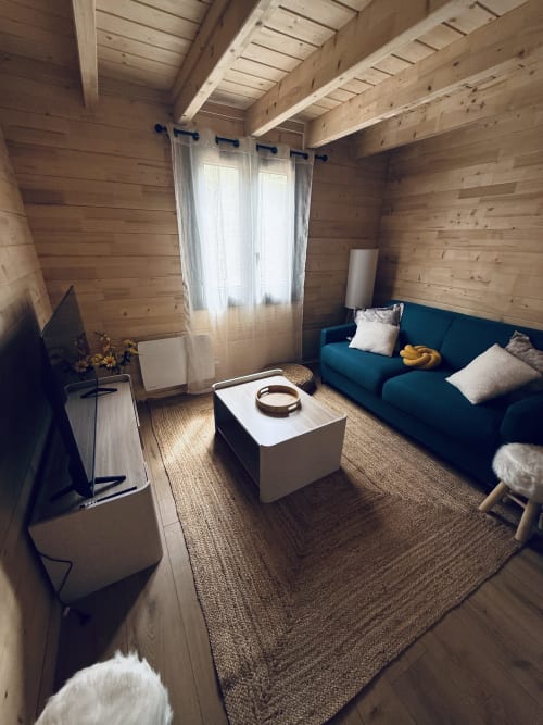 Intérieur douillet de lodge en bois avec canapé bleu, table basse blanche et grandes fenêtres donnant sur la nature
