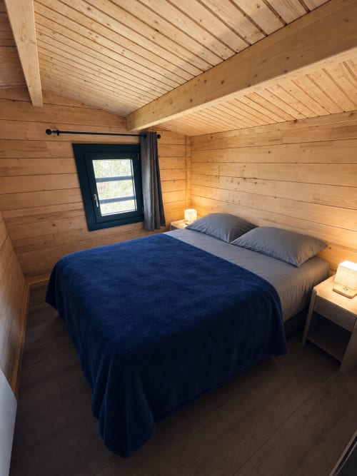 Chambre de lodge en bois douillet avec lit queen-size, couverture bleue, oreillers gris, fenêtre noire et éclairage chaleureux aux Cabanes du Belvédère