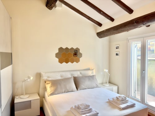 Camera da letto moderna con travi in legno a vista, biancheria bianca, arte esagonale, e porte in vetro con vista sulla città di Parma