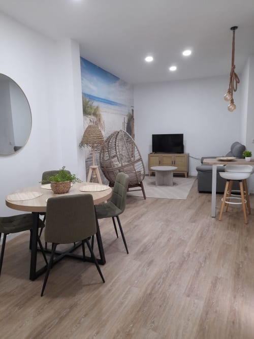 Modern apartment lounge with beach-themed mural, dining area, wicker chair, and TV at G&H Apartments Jerez