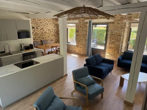 Salon ouvert à la Maison Cèdre du Clos de l'Arguenon avec cuisine, murs en pierre, fauteuils bleus et portes-fenêtres
