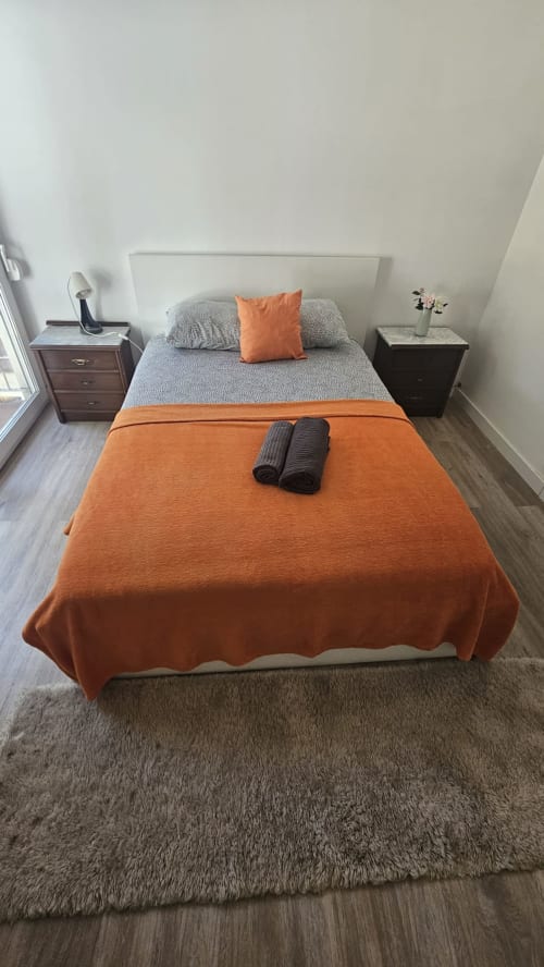Quarto moderno de hostel em Lisboa com roupa de cama laranja e cinza, cabeceira branca, mesas de cabeceira de madeira e carpete cinza.