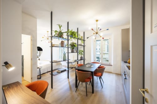 Camera d'hotel moderna a Parma con open space, libreria nera con piante, zona pranzo con sedie arancioni e lampadario sputnik