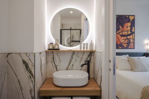 Bagno moderno dell'hotel Manu Maison di Parma con specchio illuminato rotondo, lavabo bianco a bacinella, rivestimenti in marmo e legno