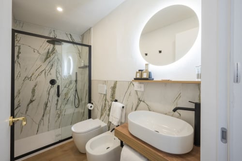 Bagno moderno a Manu Maison a Parma con piastrelle in marmo, specchio rotondo illuminato, lavabo bianco e doccia a filo