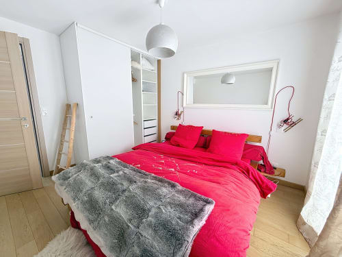 Chambre lumineuse avec literie rose vif, plaid gris, lit en bois, lampe suspendue sphérique et armoires blanches