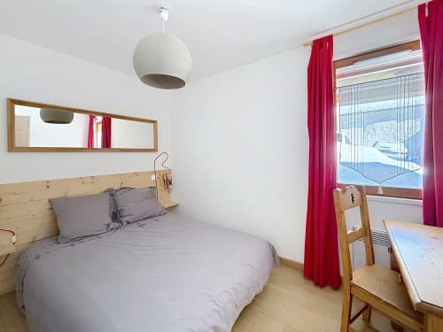 Chambre moderne aux lignes épurées avec lit en bois, parure grise et vue panoramique sur les montagnes à travers des rideaux rouges aux Tanières de Montgenèvre