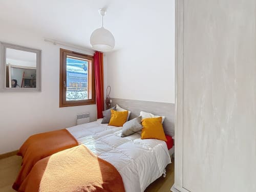 Chambre lumineuse aux draps blancs, oreillers jaunes, couverture orange, fenêtre en bois avec vue sur les montagnes aux Tanières de