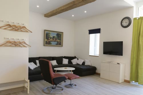 Modernes Wohnzimmer im Ferienhaus Domaine Le Piboul mit schwarzem L-förmigem Sofa, Flachbildfernseher, Holzbalken und grünen Vorhängen