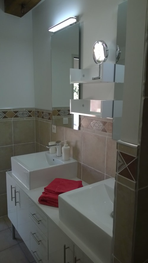 Modernes Badezimmer im Ferienhaus Les Cuves auf dem Domaine Le Piboul mit weißem Waschtisch, Waschbecken, Spiegel und roten Handtüchern