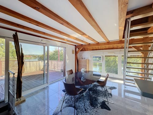 Appartement moderne au bord de l'eau à A L'ill Apart, La Wantzenau avec poutres apparentes, coin repas et vue sur l'eau depuis les portes vitrées.