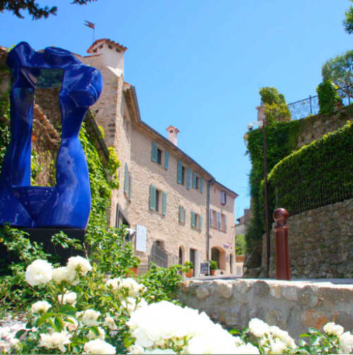 Cour des Résidences M en France avec sculpture bleue, bâtiments médiévaux en pierre couverts de lierre, fleurs blanches et ciel bleu dégagé