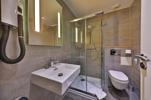 Salle de bain moderne de l'Hôtel Louvre Richelieu Paris avec douche à l'italienne, lavabo blanc, toilettes et miroir lumineux
