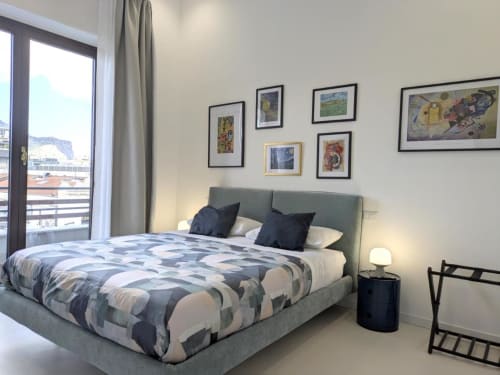 Camera moderna del BnB del Parlamento Palermo con letto rivestito grigio, copripiumino geometrico, opere d'arte incorniciate e balcone con vista città