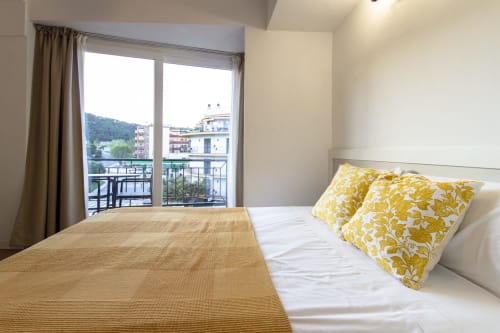 Dormitorio luminoso con cojines florales amarillos, balcón con vistas a Tossa de Mar, ropa de cama beige y paredes neutras en El Sol