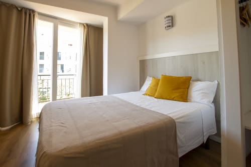 Dormitorio moderno en El Sol II Tossa de Mar con cama queen, almohadas amarillas, balcón con vista a la ciudad y luz natural