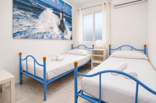 Habitación luminosa con dos camas de metal azul, ropa de cama blanca, arte de olas marinas y comodidades modernas en Living Sur, Sanlúcar de