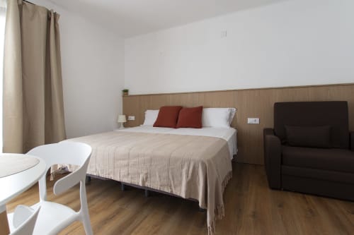 Habitación moderna del hotel El Sol II en Tossa de Mar con cama matrimonial, almohadas rojas, cabecero de madera, sillón oscuro y detalles de madera