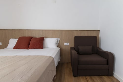 Dormitorio moderno con cabecera de madera, cama blanca, almohadas rojas y butaca marrón en El Sol II Tossa de Mar