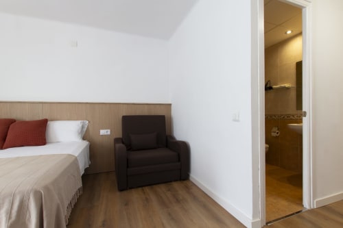 Habitación moderna del hotel El Sol II en Tossa de Mar con cama, butaca oscura, suelo de madera y baño privado