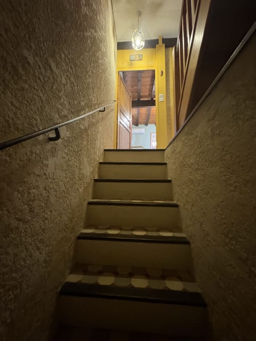Escalier étroit aux murs texturés et rampe métallique menant à une porte jaune à la Conciergerie Maurin, Arles