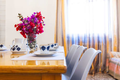 Elegante zona pranzo dell'Otranto Oasis con fiori vivaci, stoviglie decorate e tende a righe che affacciano sul balcone