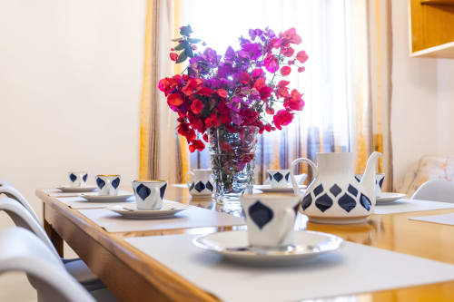 Elegante sala da pranzo dell'Oasis Apartment di Otranto con fiori rossi e viola, stoviglie decorate e finestre luminose