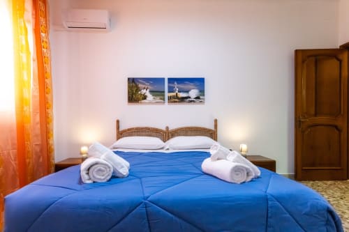 Camera luminosa all'Otranto Oasis con coperta blu, asciugamani arrotolati, letto in legno e due foto costiere incorniciate su muro bianco