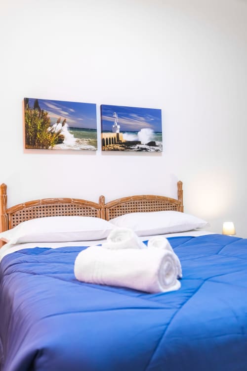 Camera Otranto Oasis con testata in legno, biancheria blu, cuscini bianchi e vedute marine costiere sopra il letto