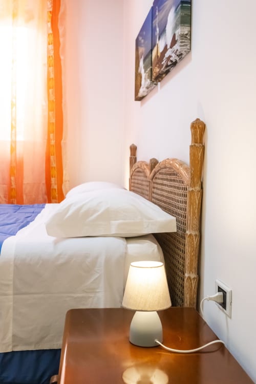 Camera da letto dell'appartamento Otranto Oasis con biancheria bianca, testata in vimini, lampada comodino e finestra con tenda arancione