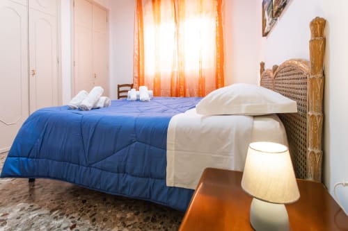 Camera luminosa all'Otranto Oasis con piumone blu, testata in vimini, cuscini bianchi, tende arancioni e comodino in legno