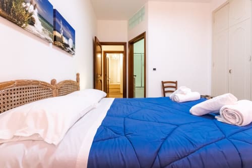 Camera da letto dell'appartamento Otranto Oasis con coperta trapuntata blu, testata in legno, cuscini bianchi e asciugamani arrotolati