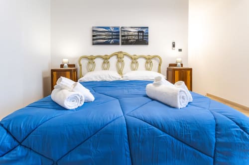 Camera spaziosa all'Otranto Oasis con piumino blu, asciugamani bianchi arrotolati, testata dorata ornamentale e vista sul paesaggio costiero