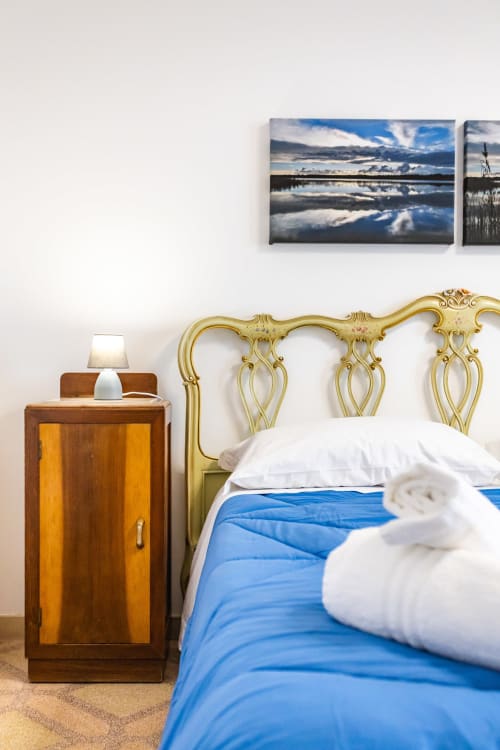 Camera elegante dell'Otranto Oasis con testata in ottone ornato, biancheria blu, comodino in legno e opera paesaggistica
