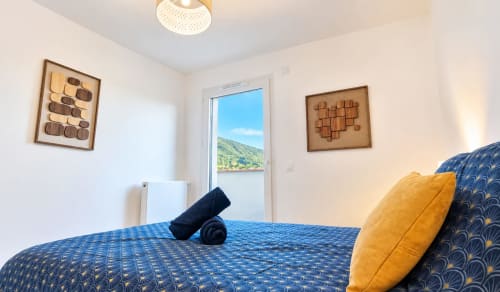 Chambre lumineuse chez NN Patrimoine à Douvaine avec lit à motifs bleus, coussin jaune et vue sur le lac de montagne depuis la porte-fenêtre