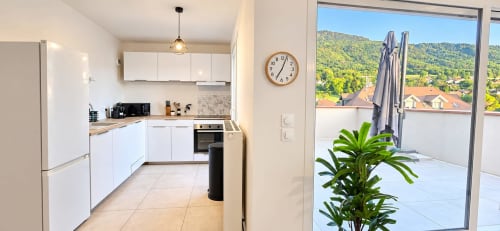Cuisine blanche moderne avec comptoirs en bois, cuisinière et grand balcon surplombant les collines verdoyantes et toits du village de Douvaine