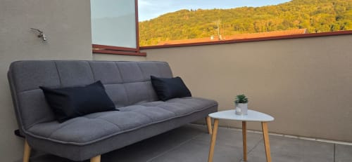 Canapé gris moderne avec coussins noirs sur le balcon de l'appartement L'Idéal, vue sur les collines boisées de Douvaine