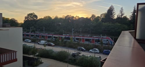 Train rouge arrêté en gare de Douvaine avec voitures garées et arbres au coucher du soleil, vue du balcon de l'appartement NN Patrimoine