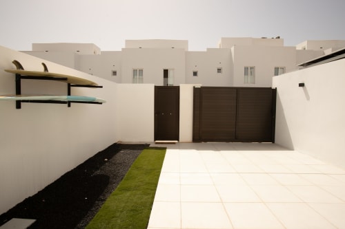 Patio de Villa MERAKI con muros blancos, puerta de madera oscura, jardín verde y terraza moderna en Corralejo, Fuerteventura