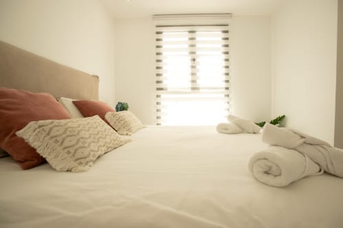 Dormitorio luminoso en Villa MERAKI en Corralejo con ropa de cama neutra, almohadas de terracota y persianas rayadas que dejan pasar la luz natural