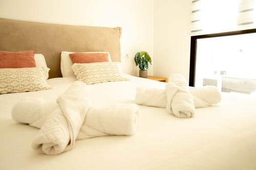 Dormitorio luminoso en Villa MERAKI, Fuerteventura, con cama, toallas enrolladas, almohadas coral, planta en maceta y balcón con vistas al mar