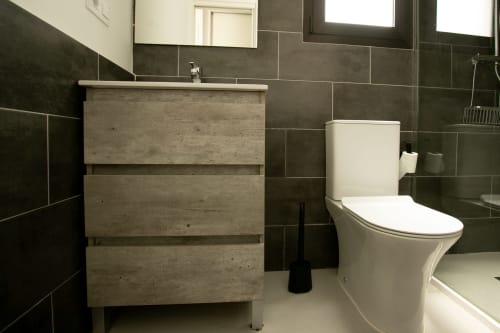 Baño moderno en Villa MERAKI con tocador de madera, inodoro blanco y paredes de azulejos oscuros en Gozamena, Fuerteventura
