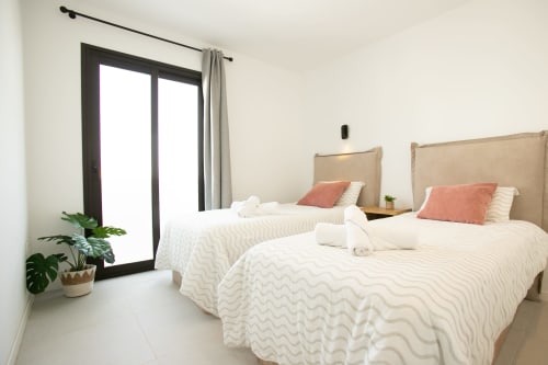 Luminosa habitación en Villa MERAKI con dos camas, ropa de cama crema, almohadas coral, cabeceros beige y acceso a balcón con vistas al mar