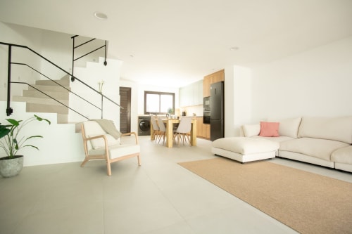 Espacio de sala moderno con sofá crema, mesa de comedor de madera, cocina y escalera con barandillas metálicas en Fuerteventura