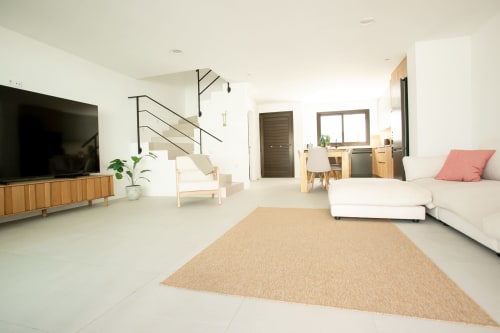 Salón Villa MERAKI Corralejo con vista al mar, sofá moderno en forma de L, mueble televisivo en madera, alfombra beige, escaleras y cocina abierta