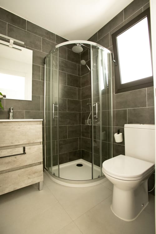 Baño moderno con ducha de azulejos oscuros, inodoro blanco y tocador de madera clara en Villa MERAKI en Gozamena