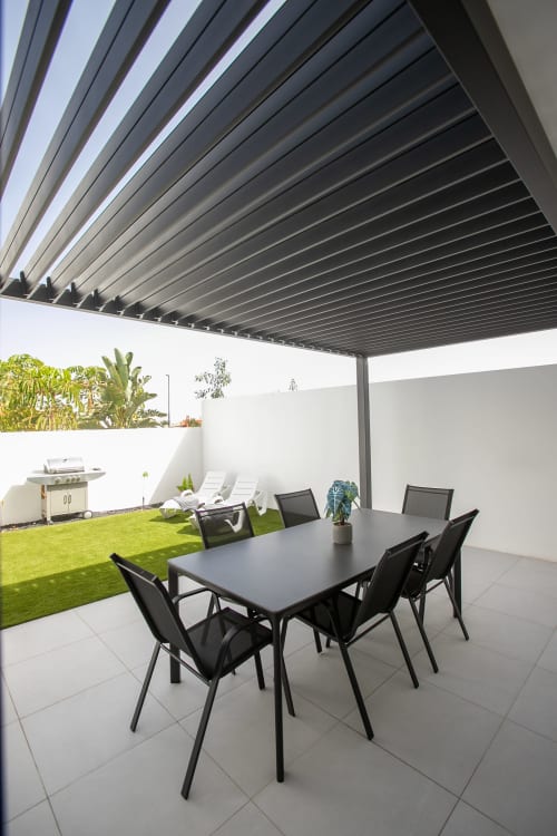 Patio moderno de Villa MERAKI con mesa de comedor, sillas negras, pérgola orientable y jardín cuidado en Fuerteventura