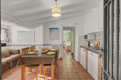 Comedor y cocina luminosos en Can Gat Vell, Girona, con suelos de terracota, mesa de madera, armarios blancos y estilo costero